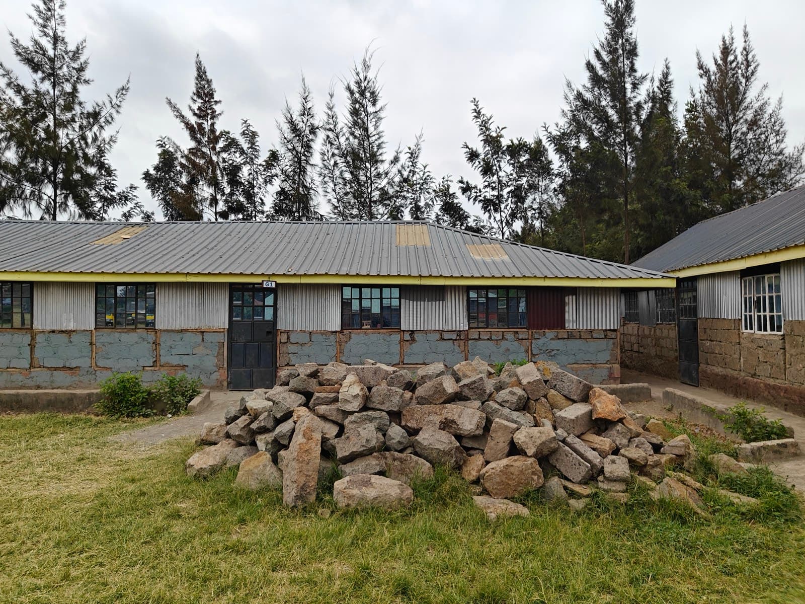 A long semi-permanent classroom block, stone to the window sills and iron sheet above, under a pitched roof with yellow fascia boards