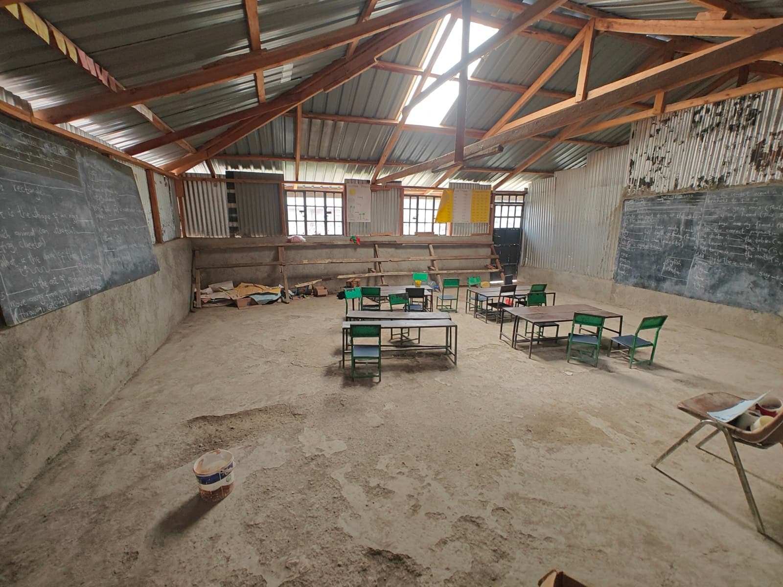 A large empty classroom with a concrete floor, green metal desks, and blackboards on two walls under exposed timber trusses