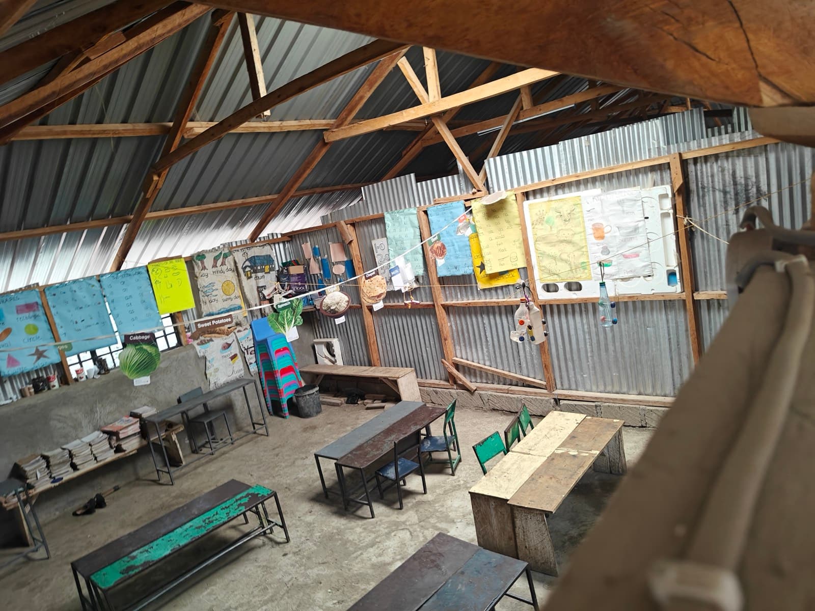 A classroom of iron sheets and timber roof trusses, charts pinned along the walls above metal desks