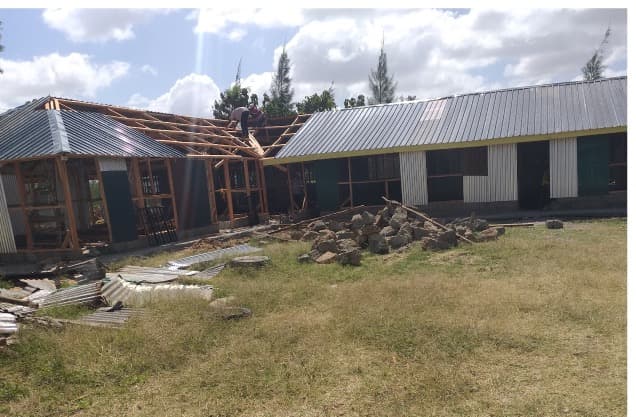 A man on the roof timbers of a half-built classroom block, iron sheets stacked on the grass below