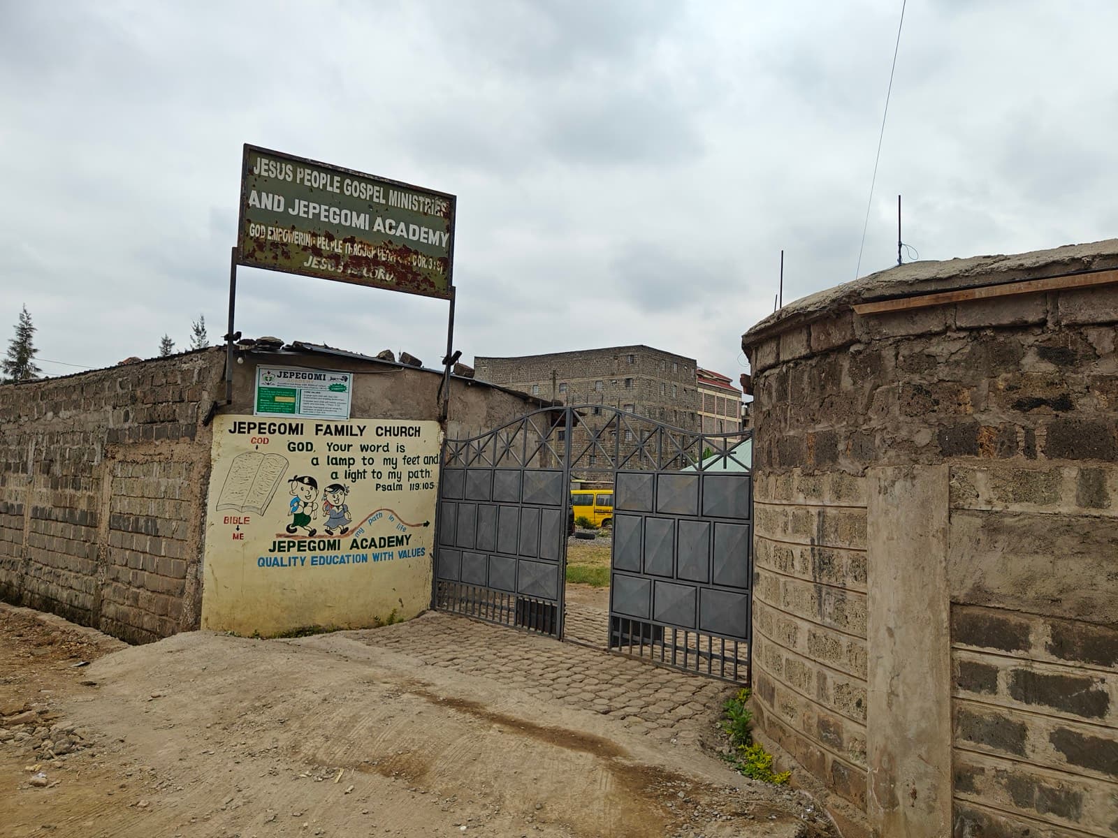 The school gate from the road, under a painted board reading Jesus People Gospel Ministries and Jepegomi Academy