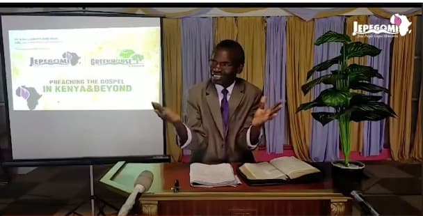 Pastor Simon Nderitu preaching from a lectern with an open Bible, a projector screen beside him reading Preaching the Gospel in Kenya and Beyond