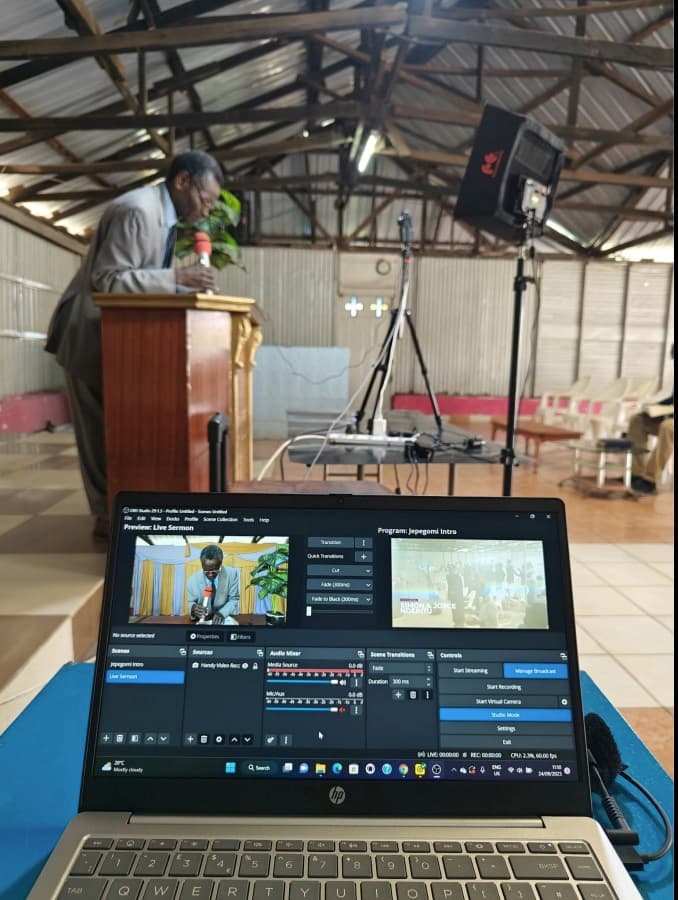 A laptop running OBS Studio on a stand at the back of the church, its screen showing the live sermon being mixed, while Pastor Simon preaches from the pulpit behind it