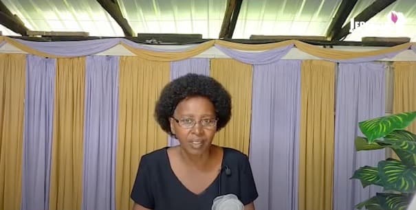Joyce Nderitu speaking to camera in front of draped yellow and lilac curtains