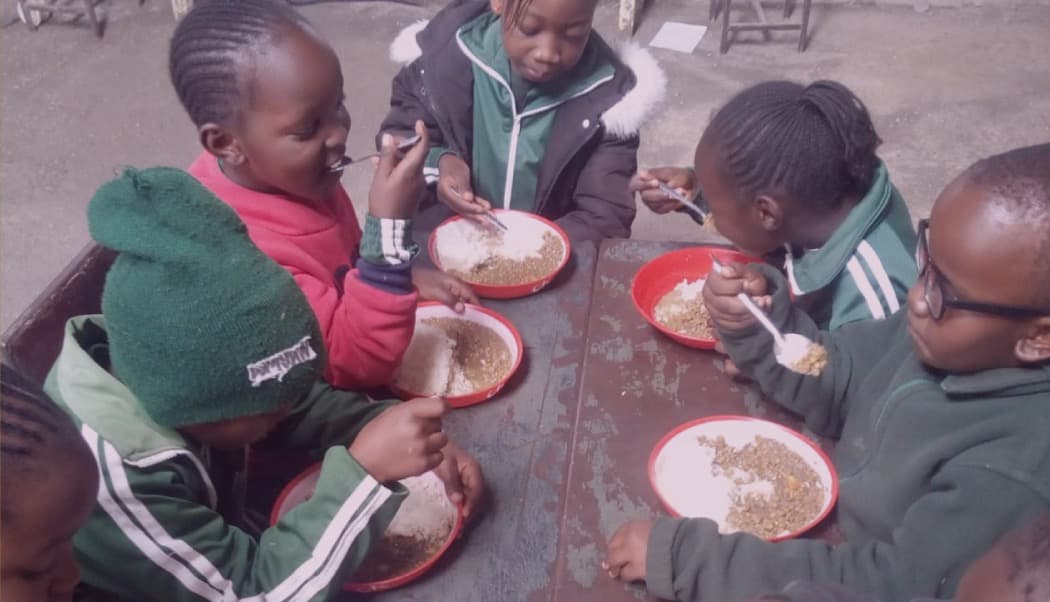 Five children in green uniform eating rice and beans from red plates at a wooden desk