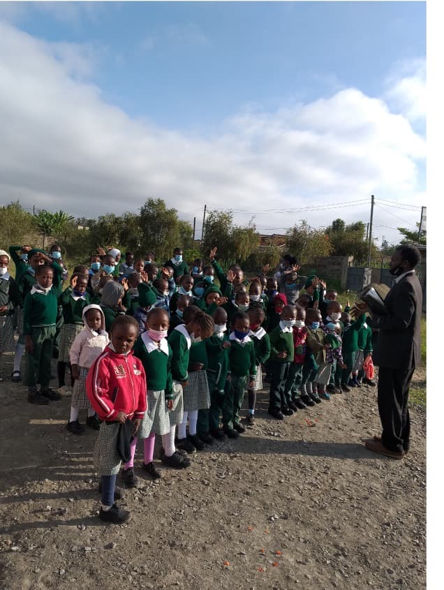 A long line of children in green uniform standing outdoors at assembly, a teacher reading to them
