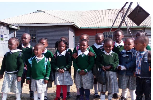 A group of young children in green jumpers and checked uniform standing together outside the school