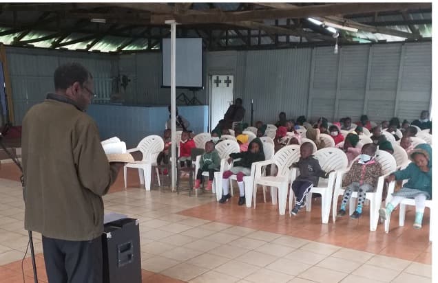 A man standing at a lectern reading to rows of seated children