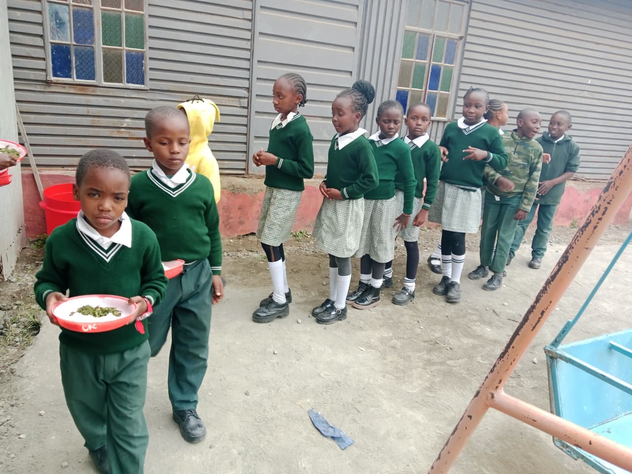Children in green uniform queueing along a wall, plates in hand
